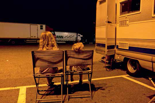 Zwei Kinder von hinten auf Campingstühlen auf einem Autobahnrastplatz, es ist Nacht.