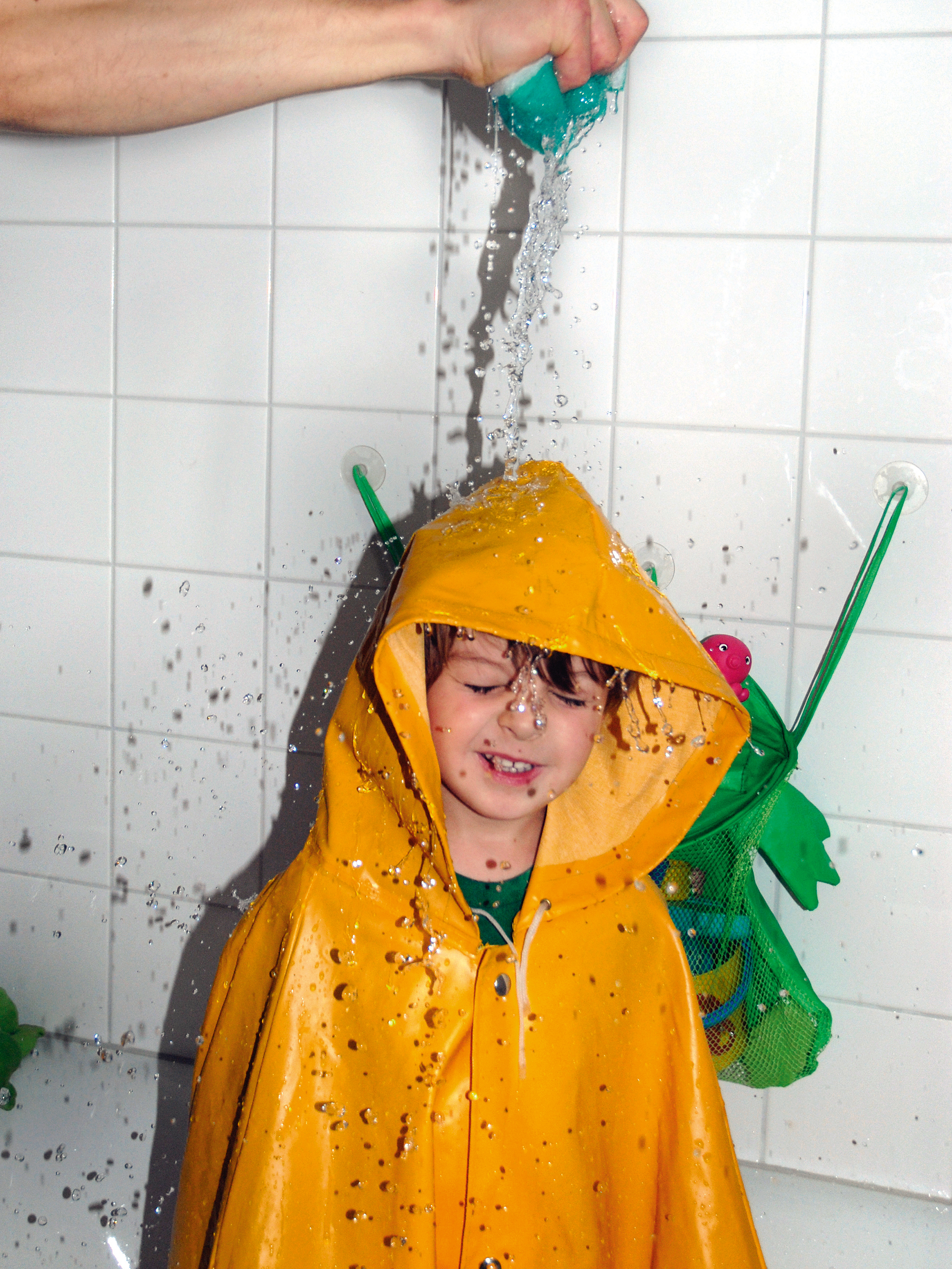 Kind mit Regenmantel in der Badewanne wird von den Eltern nass gemacht
