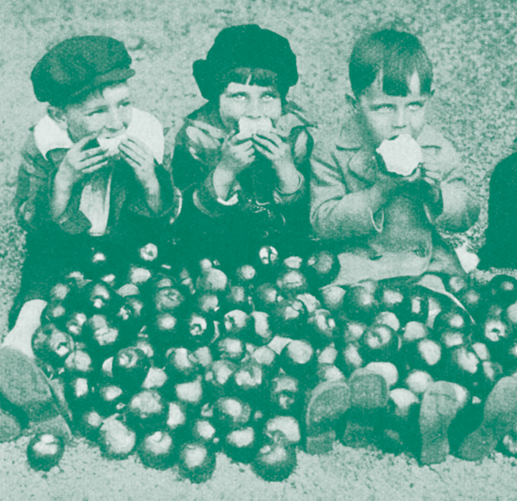 Drei Kinder sitzen am Boden und essen Äpfel. Ihre Beine sind von Äpfeln bedeckt.