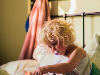 Kind mit Schnuller sitzt auf einem Bett und bedient ein Tablet